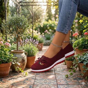 May include: Burgundy slip-on shoes with a white sole and red trim. The shoes have a bow detail with a small embellishment. The image shows the shoes in an outdoor setting with potted plants and flowers.