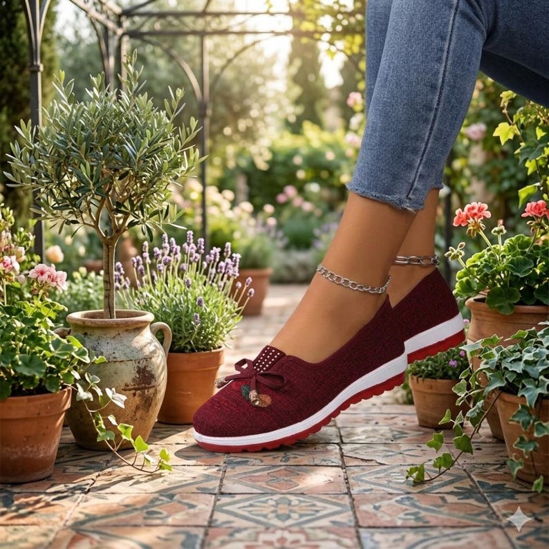 May include: Burgundy slip-on shoes with a white sole and red trim. The shoes have a bow detail with a small embellishment. The image shows the shoes in an outdoor setting with potted plants and flowers.