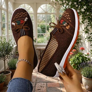 May include: Brown slip-on shoes with decorative floral accents and a bow. The shoes have a white sole with an orange stripe and a breathable mesh interior. The shoes are designed for casual wear.