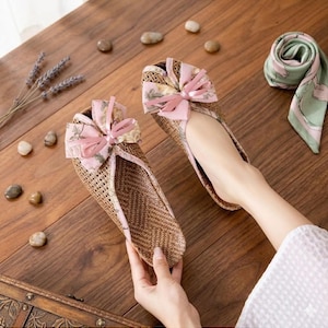 May include: A pair of woven, open-toe flats with pink floral bows. The shoes have a natural, light brown color and are decorated with a pink floral pattern. The flats are designed for comfort and style.