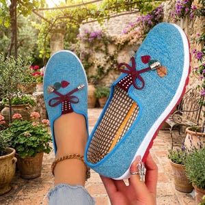 May include: Light blue slip-on shoes with decorative accents. The shoes feature a white sole with a red trim, a blue trim, and a decorative bow. The interior has a brown and white checkered pattern. The shoes are designed for casual wear.