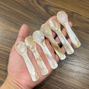 Puede incluir: Un juego de seis cucharas pequeñas, nacaradas, con un brillo natural e iridiscente. Las cucharas tienen un cuenco redondeado y un mango delgado, con una combinación de colores blanco crema y dorado. Las cucharas se sostienen en una mano.