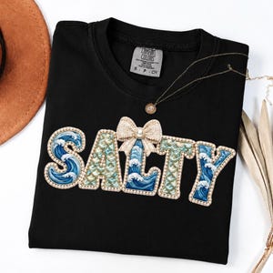 May include: Black t-shirt with the word "SALTY" embroidered in a whimsical font. The letters feature a wave design in shades of blue and teal, accented with a pearl-like trim and a bow. A gold necklace is draped across the shirt.