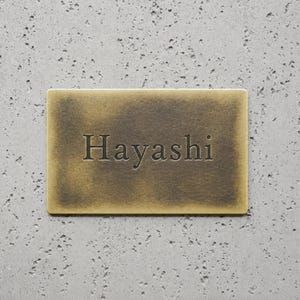 May include: A rectangular brass plaque with a brushed, aged finish featuring the word Hayashi engraved in the center in a dark, serif font. The metal surface displays subtle variations in tone, ranging from light gold to deep bronze.