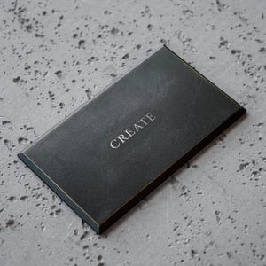 May include: A rectangular black plaque with beveled edges and a thin gold-colored border. The word CREATE is embossed in the center in a serif font. The surface of the plaque has a matte, slightly textured finish, providing a clean look.