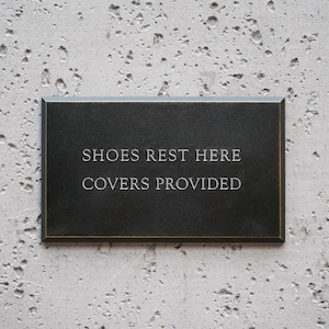 May include: A rectangular black sign with a thin gold border featuring the engraved white text SHOES REST HERE COVERS PROVIDED. The sign has a smooth, dark surface and is designed for clear communication in a home or professional setting.