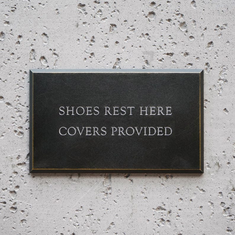 May include: A rectangular black sign with a thin gold border featuring the engraved white text SHOES REST HERE COVERS PROVIDED. The sign has a smooth, dark surface and is designed for clear communication in a home or professional setting.