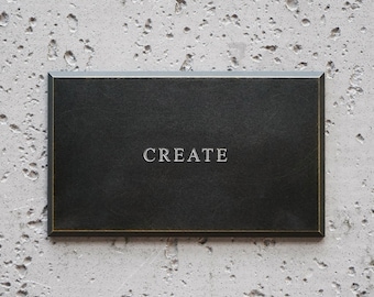 May include: A rectangular black plaque with beveled edges and a thin gold-toned border. The word CREATE is centered on the surface in a serif font. The plaque has a smooth, matte finish and a clean, structured design with sharp corners.