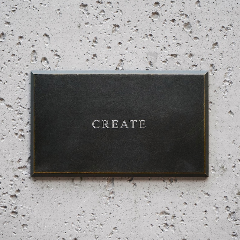 May include: A rectangular black plaque with beveled edges and a thin gold-toned border. The word CREATE is centered on the surface in a serif font. The plaque has a smooth, matte finish and a clean, structured design with sharp corners.