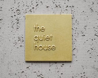 May include: A square, gold-colored metal sign with the words "the quiet house" embossed in a serif font. The sign is mounted on a textured, gray concrete wall. The sign is a decorative element.