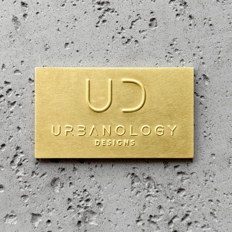 May include: A rectangular gold-colored metal plaque featuring an embossed logo with the letters UD at the top and the text URBANOLOGY DESIGNS printed in a sans-serif font below. The surface of the plaque has a smooth, matte metallic finish.