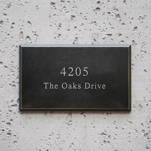 May include: A rectangular black house sign with a thin gold border. The sign features the engraved white text 4205 on the top line and The Oaks Drive on the bottom line, centered in a clean, serif font against a smooth, dark surface.