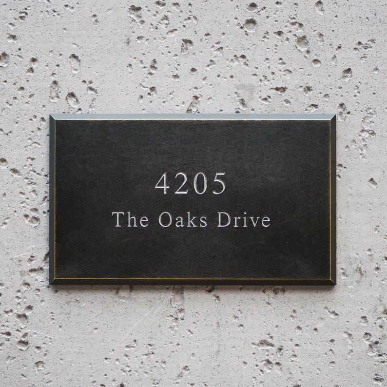 May include: A rectangular black house sign with a thin gold border. The sign features the engraved white text 4205 on the top line and The Oaks Drive on the bottom line, centered in a clean, serif font against a smooth, dark surface.
