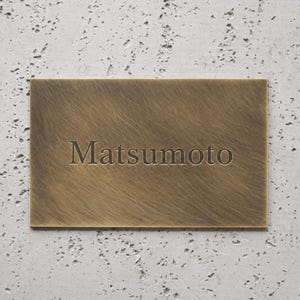 May include: A rectangular, bronze-colored metal sign with the name "Matsumoto" engraved in a sans-serif font. The sign has a textured surface and is mounted on a gray, speckled concrete wall.