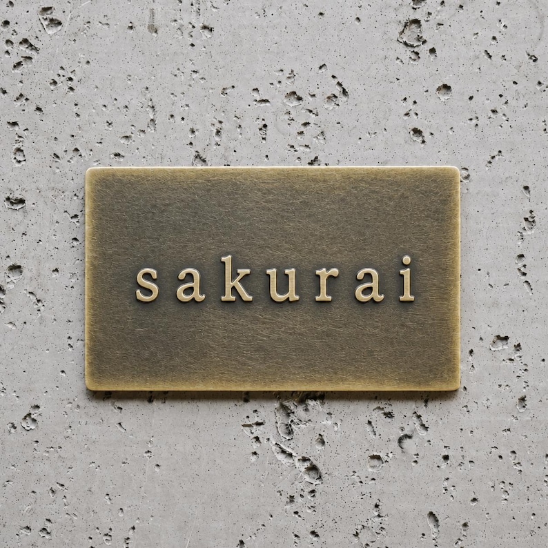 May include: A rectangular brass plaque with a brushed metallic finish featuring the word "sakurai" embossed in lowercase serif lettering centered on the surface. The plaque has a smooth, flat face with clean edges and a warm, aged gold tone.