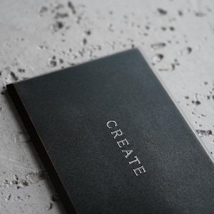 May include: A rectangular black notebook with a textured cover featuring the word CREATE embossed in silver capital letters centered on the front. The notebook has a clean, dark aesthetic with a matte finish and sharp, defined edges.