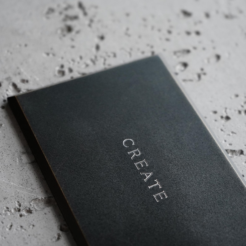 May include: A rectangular black notebook with a textured cover featuring the word CREATE embossed in silver capital letters centered on the front. The notebook has a clean, dark aesthetic with a matte finish and sharp, defined edges.