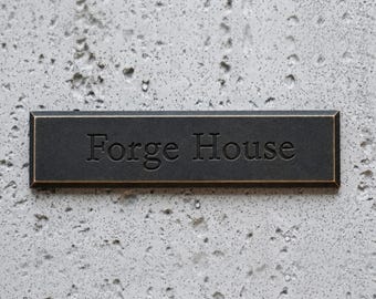 May include: A rectangular, dark metal sign with the embossed text "Forge House" in a serif font. The sign has a bronze-colored border and is mounted on a textured, light gray concrete wall.