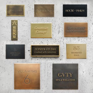 May include: A collection of rectangular and square metal signs with embossed text. The signs are in various colors, including gold, bronze, and black. Text includes "castanca", "Cafe de la Paix", "HOUSE of PSALM", "Greatcoat Cottage", and "ZEPHYR STUDIO".