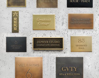May include: A collection of rectangular and square metal signs with embossed text. The signs are in various colors, including gold, bronze, and black. Text includes "castanca", "Cafe de la Paix", "HOUSE of PSALM", "Greatcoat Cottage", and "ZEPHYR STUDIO".