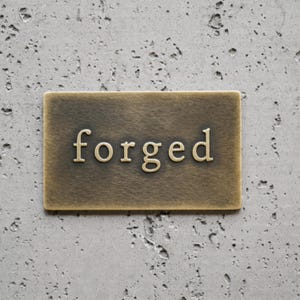 May include: A rectangular brass plaque with a brushed, antiqued finish featuring the word forged in raised, serif-style lettering centered on the surface. The metal plate has a smooth, metallic texture with visible signs of aging and oxidation.
