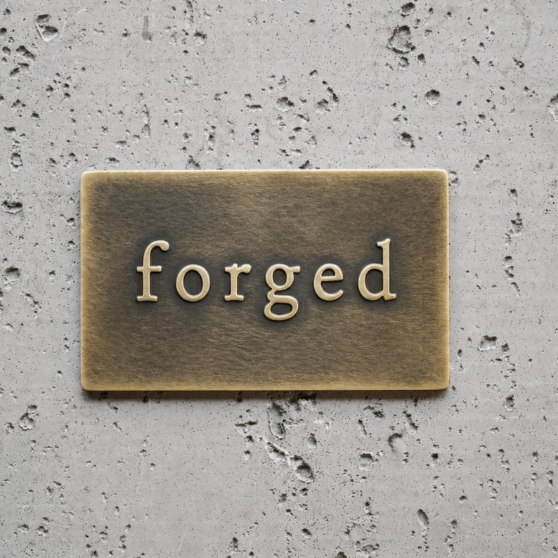 May include: A rectangular brass plaque with a brushed, antiqued finish featuring the word forged in raised, serif-style lettering centered on the surface. The metal plate has a smooth, metallic texture with visible signs of aging and oxidation.