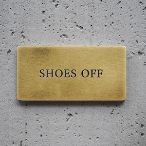May include: A rectangular brass sign with a brushed metallic finish featuring the text SHOES OFF engraved in black capital letters. The sign has a smooth, flat surface and clean, straight edges, presenting a clear and legible instructional message.