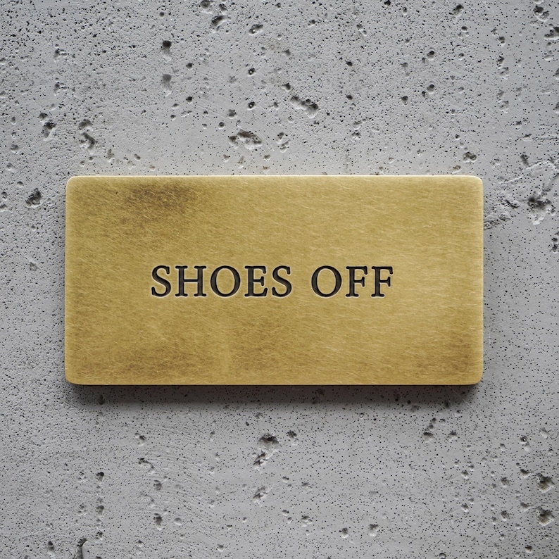 May include: A rectangular brass sign with a brushed metallic finish featuring the text SHOES OFF engraved in black capital letters. The sign has a smooth, flat surface and clean, straight edges, presenting a clear and legible instructional message.