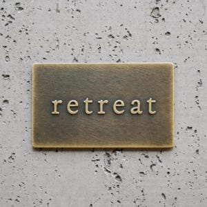 May include: A rectangular brass-toned metal sign with a brushed finish featuring the word retreat embossed in the center in a serif font. The sign has a flat surface and clean edges, presenting a metallic appearance with subtle color variations.