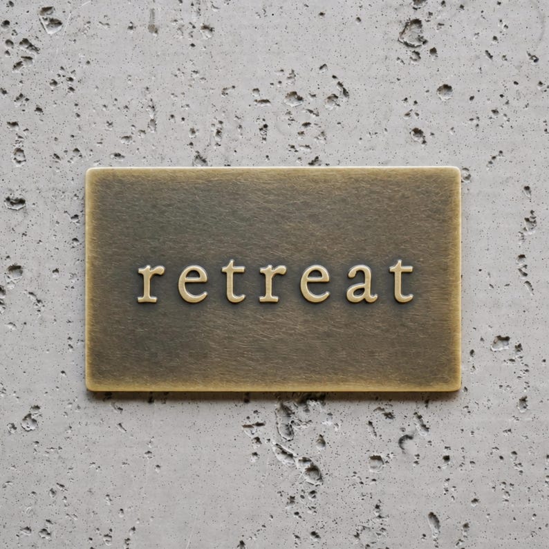 May include: A rectangular brass-toned metal sign with a brushed finish featuring the word retreat embossed in the center in a serif font. The sign has a flat surface and clean edges, presenting a metallic appearance with subtle color variations.