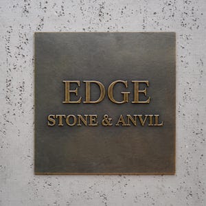 May include: A square, dark bronze-colored metal plaque featuring the raised, metallic gold-toned text "EDGE" on the top line and "STONE & ANVIL" on the bottom line. The plaque has a smooth, flat surface with a subtle metallic sheen and clean edges.