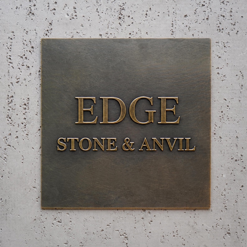 May include: A square, dark bronze-colored metal plaque featuring the raised, metallic gold-toned text "EDGE" on the top line and "STONE & ANVIL" on the bottom line. The plaque has a smooth, flat surface with a subtle metallic sheen and clean edges.