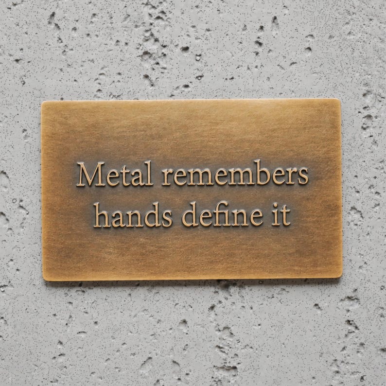 May include: A rectangular bronze-colored metal plaque with a brushed finish. The text "Metal remembers hands define it" is embossed in a serif font across the center of the plaque, featuring dark lettering that contrasts with the metallic surface.