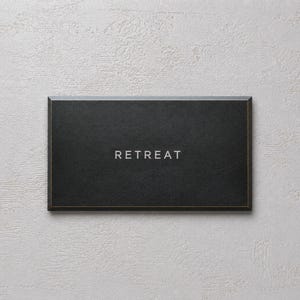May include: A rectangular black sign with a thin gold border and the word RETREAT printed in white capital letters centered on the front surface. The sign has a smooth, matte finish and a clean, simple design with sharp, defined edges.