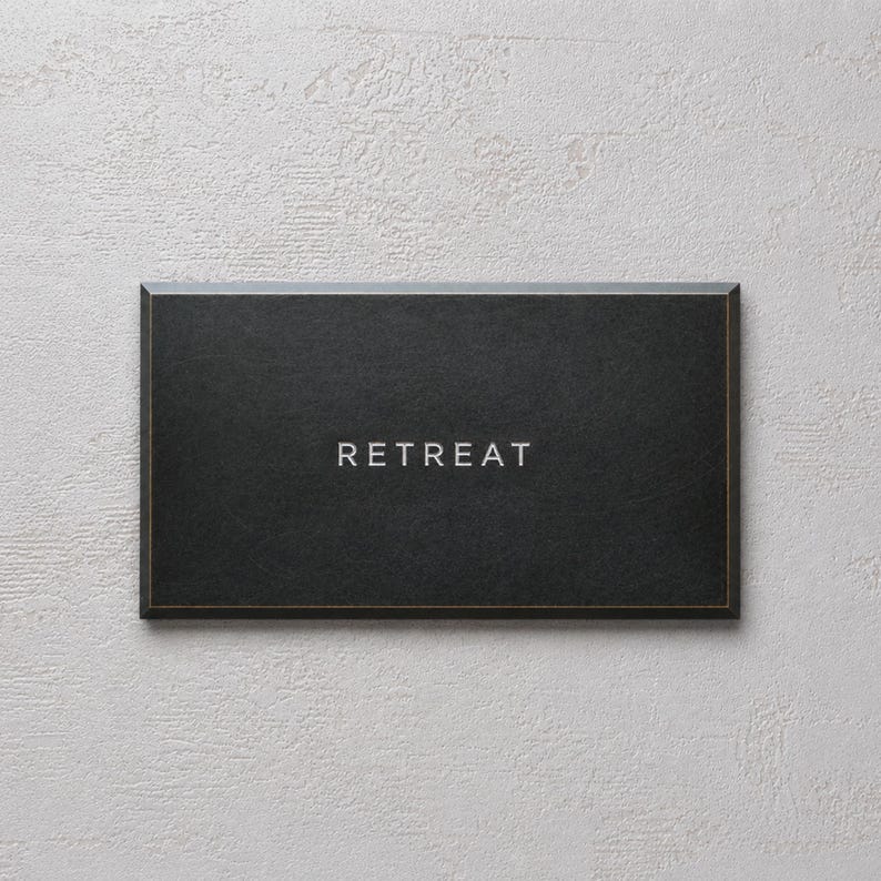 May include: A rectangular black sign with a thin gold border and the word RETREAT printed in white capital letters centered on the front surface. The sign has a smooth, matte finish and a clean, simple design with sharp, defined edges.