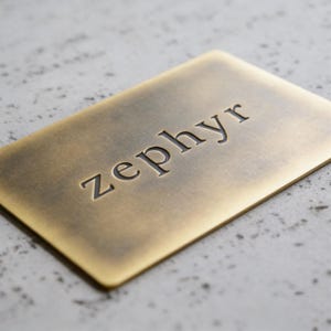 May include: A rectangular brass-colored metal plate with a brushed finish. The word zephyr is engraved in lowercase, sans-serif letters across the center of the plate. The metal has a slightly darkened, aged patina around the edges and lettering.