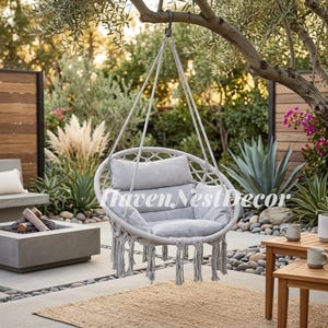 May include: A round, light gray macrame hanging chair suspended from a tree branch. The chair features a woven rope frame, a soft padded seat cushion, a matching backrest pillow, and decorative fringe tassels hanging from the bottom edge.