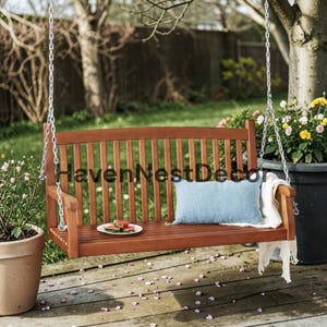 May include: A wooden porch swing with a slatted back and seat, suspended by silver metal chains. A light blue rectangular pillow and a white throw blanket rest on the seat. A small white plate with watermelon slices sits on the wooden slats.
