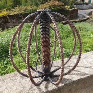 Puede incluir: Una escultura esférica construida con alambre de metal oscuro trenzado. El objeto presenta un cilindro central vertical rodeado por múltiples anillos circulares de alambre que se cruzan, todos con una apariencia texturizada y trenzada en tonos bronce y negro.