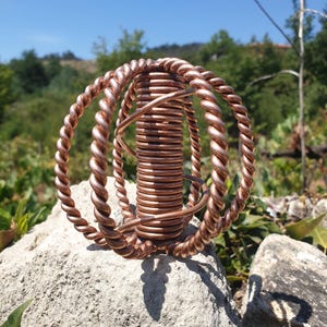 Peut inclure: Une sculpture en fil de cuivre composée d'une bobine verticale centrale entourée de trois anneaux concentriques en fil de cuivre torsadé. L'objet métallique présente une finition brun rougeâtre et est formé de plusieurs brins de fil enroulés.