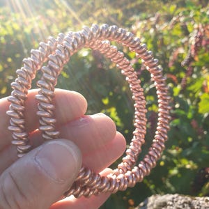 Może przedstawiać: Dwie spiralne gumki do włosów w kolorze różowego złota z metalicznym wykończeniem. Gumki mają ciągły, zwinięty wzór tworzący okrągły kształt, co zapewnia elastyczną i rozciągliwą strukturę do upinania włosów w różne fryzury.
