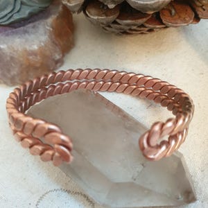 Può includere: Un bracciale a fascia in rame caratterizzato da un design a doppio filo a corda intrecciata. Il bracciale è aperto e presenta una finitura metallica color marrone rossastro con un motivo uniforme e ripetitivo lungo le due fasce metalliche.