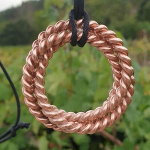 May include: A circular pendant crafted from two intertwined strands of polished copper wire, forming a twisted rope texture. The metallic ring is suspended from a simple black cord, showcasing a warm, reflective finish and a symmetrical design.