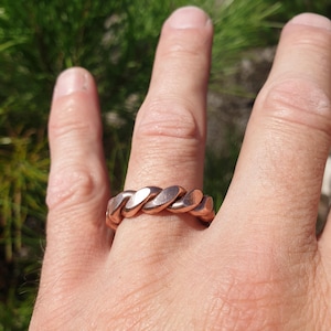 Könnte beinhalten: Ein metallisch kupferfarbener Ring mit einem durchgehenden gedrehten Seil- oder Kabeldesign. Das Band hat eine polierte Oberfläche mit ausgeprägten diagonalen Segmenten, die sich um den gesamten Umfang des kreisförmigen Schmuckstücks ziehen.