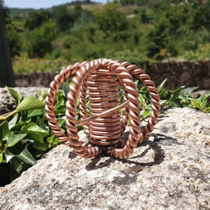 Peut inclure: Une sculpture sphérique fabriquée en fil de cuivre poli, composée d'une bobine centrale étroitement enroulée entourée de trois anneaux entrelacés faits de brins de cuivre torsadés, formant un objet métallique géométrique et symétrique.