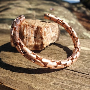 Pode incluir: Uma pulseira de cobre tipo bracelete com um padrão repetitivo de elos entrelaçados e acabamento metálico polido. A pulseira tem um design aberto, exibindo uma série de segmentos arredondados e texturizados que formam uma estrutura contínua.