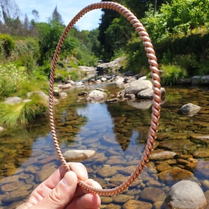 May include: A circular torque necklace crafted from a single strand of twisted copper wire. The metal has a warm, metallic reddish-brown hue and a continuous spiral texture that runs along the entire length of the circular band.