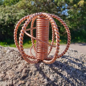 Könnte beinhalten: Eine Kupferskulptur mit einer zentralen Spule, umgeben von gedrehten Drahtringen. Die Skulptur steht auf einem strukturierten Felsen, mit einem verschwommenen grünen Hintergrund aus Bäumen und Himmel. Das Kupfer hat einen warmen, rötlich-braunen Farbton.