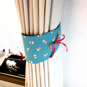 以下が含まれることがあります： 黒い羽を持つ小さな黄色い蜂の繰り返し模様が特徴のティール色のカーテンタッセル。布製のバンドは、側面に結ばれた赤とピンクの編み込みコードでベージュのカーテンの周りに固定されています。