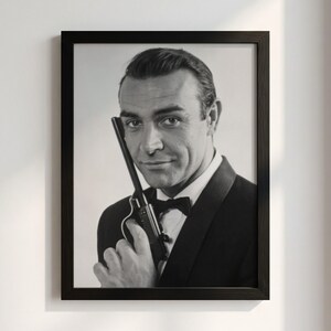 May include: A black and white framed portrait of a man wearing a tuxedo with a bow tie, holding a pistol near his face. The man has short dark hair and is looking directly at the viewer. The frame is black with a simple, clean design.